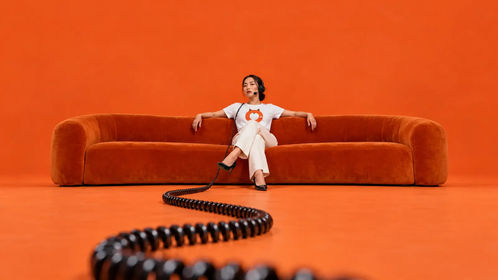 A Thai telesales agent in a headset sits on an orange sofa, a coiled cord leading to her